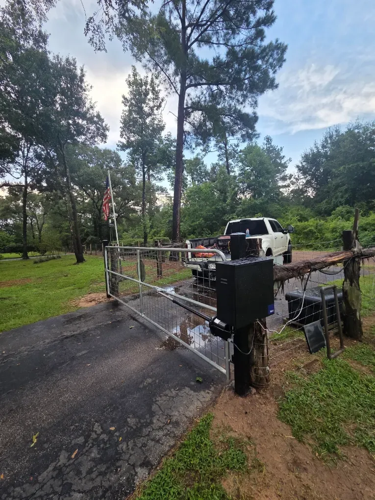 Pipe and wire ranch gate with a swing operator on a wooden post, wooded acreage north of Houston